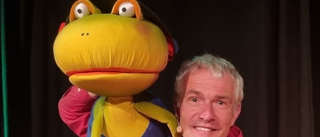 Ein Mann in einem roten Hemd hält eine bunte Puppe mit großen Augen und einem Hut und lächelt für ein Foto.