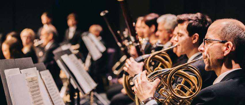 Eine Gruppe von Musikern tritt bei einem Konzert auf, wobei der Fokus auf einem Mann liegt, der das Waldhorn spielt. Die Musiker sitzen in einer Reihe, mit Notenständern vor ihnen.