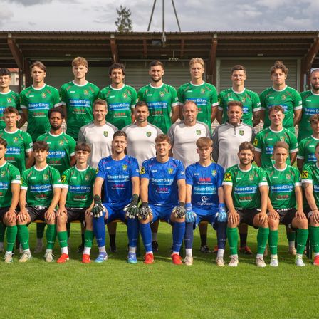 Die Fußballmannschaft in grünen Trikots posiert für ein Mannschaftsfoto auf dem Feld mit einem Gebäude im Hintergrund.