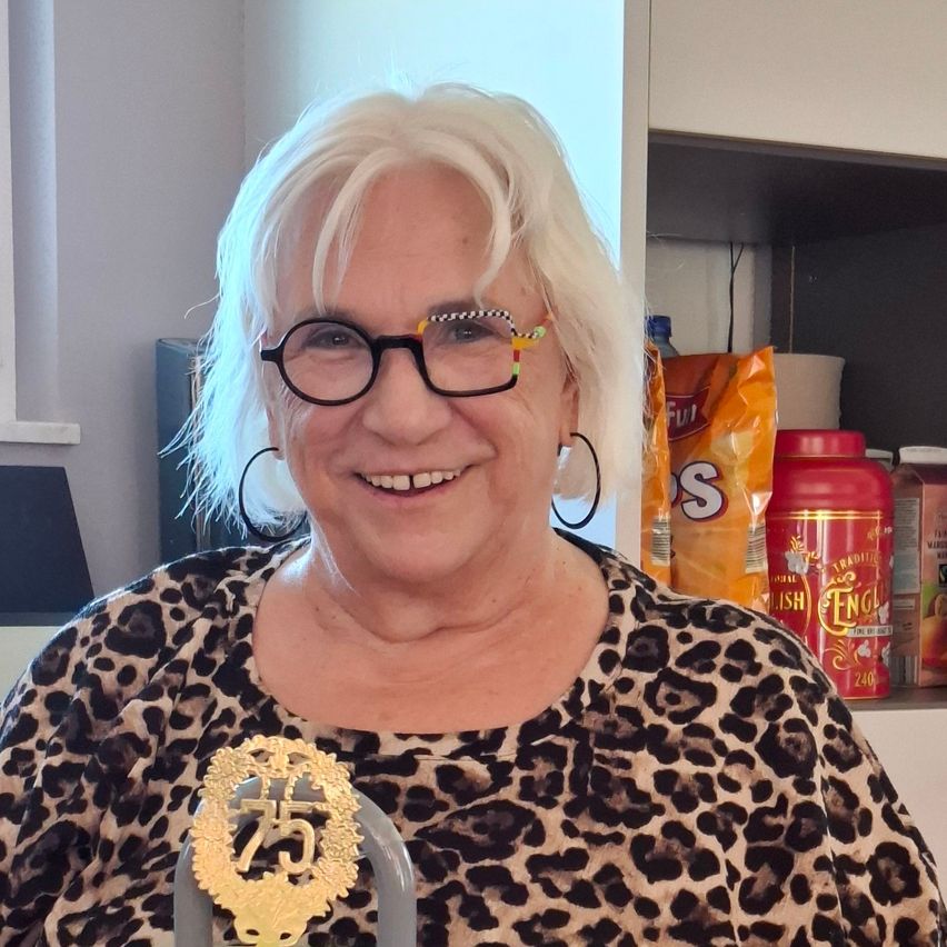 Eine Frau im Leopardenmusterhemd mit Brille posiert für ein Foto. Hinter ihr steht ein Regal mit Snacks und Getränken. Sie hält eine goldene Auszeichnung mit 25 darauf.
