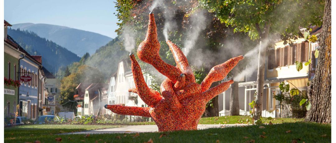 Eine farbenfrohe Skulptur mit mehreren Auslässen, die Wassernebel emittieren, steht in einem Park, umgeben von üppigem Baumbestand und Gebäuden im Hintergrund.