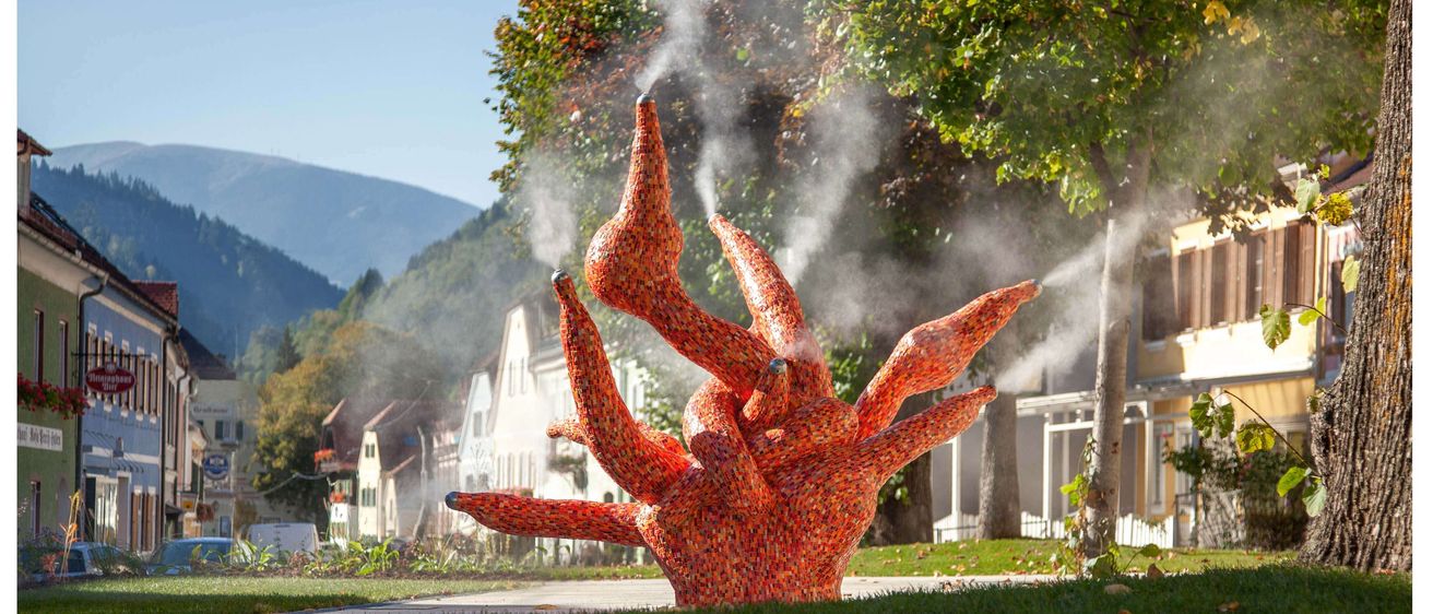 Eine bunte, farbenfrohe Skulptur steht in einem Park und gibt Dampf aus ihren Armen ab. Dahinter sind Gebäude mit Bergen in der Ferne zu sehen.