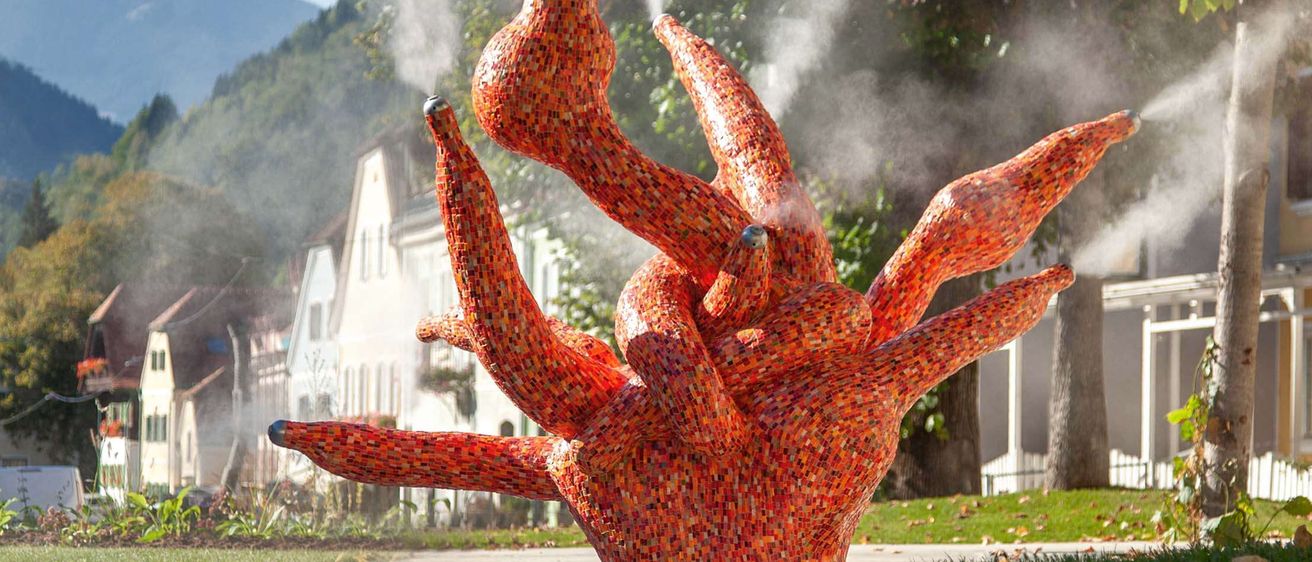 Eine farbenfrohe, komplizierte Skulptur aus Fliesen steht in einem Park, mit Nebel, der aus der Spitze austritt. Dahinter befinden sich Gebäude und Bäume, sowie Berge in der Ferne.