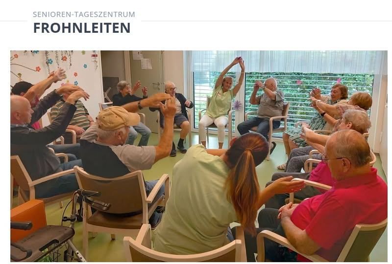 Eine Gruppe älterer Menschen nimmt in einem gut beleuchteten Raum an einer Aktivität teil, in lässiger Kleidung. Einige sitzen, andere stehen und beteiligen sich an Übungen oder Aktivitäten. Der Raum hat Fenster mit Vorhängen, und ein Rollstuhl ist vorhanden.