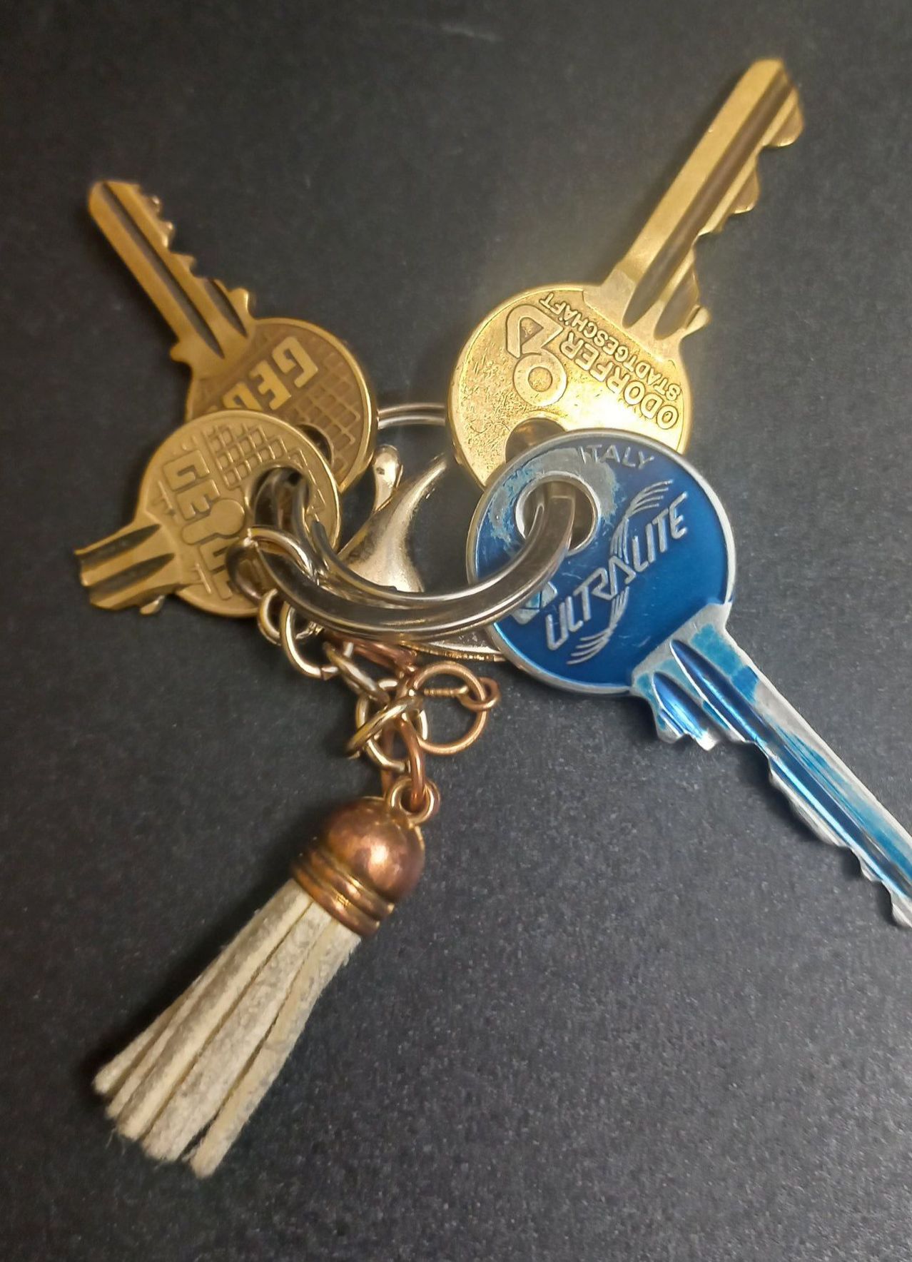 Eine Reihe verschiedener Schlüssel, einschließlich Ultralite, Italien und anderer, auf einer schwarzen Oberfläche mit Quaste und Kette.