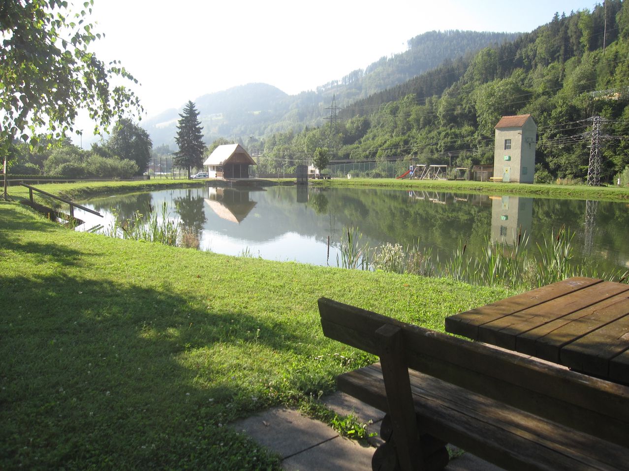 Ein ruhiger Teich mit einem Holzhaus am Ufer, umgeben von üppigem Grün und Bergen im Hintergrund.