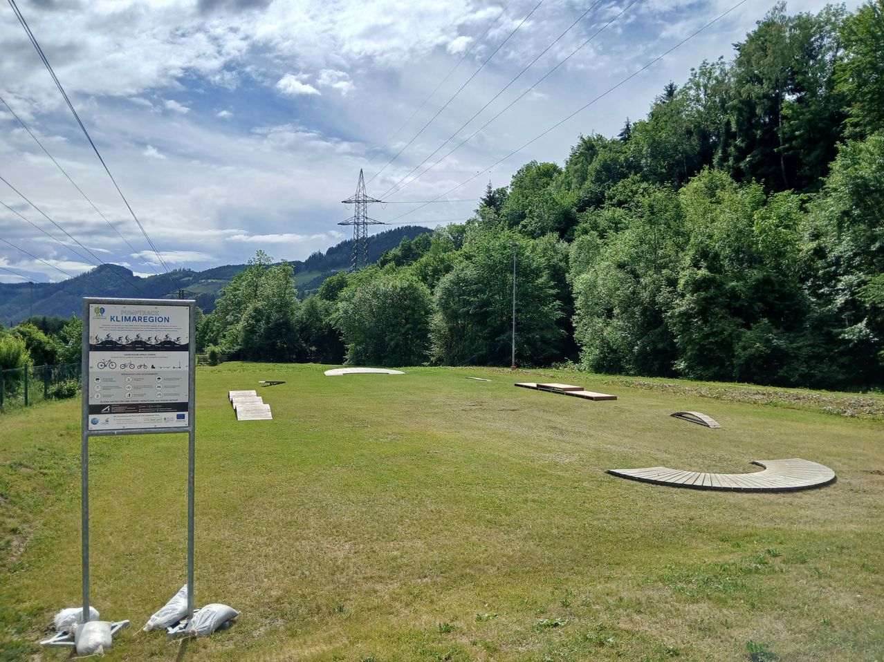 An open grassy field with a signboard labeled Klimaregion. The field has wooden ramps and a tower is visible in the background.