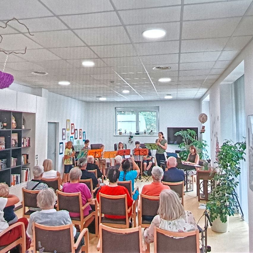 A group of people are gathered in a room, listening to a small band playing music. The band is positioned on a stage, and the audience is seated in chairs. The room has a bookshelf, a window, and a plant.
