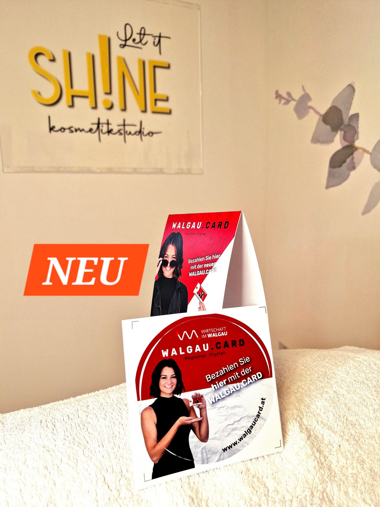 Eine Werbeanzeige für die Walgau Card ist auf einer weißen Oberfläche platziert. Sie zeigt eine Frau, die eine Karte hält, und den Text 'Bezahlen mit der Walgau.Card'. Der Hintergrund umfasst eine Wand mit einer Pflanze und dem Wort 'Neu'.