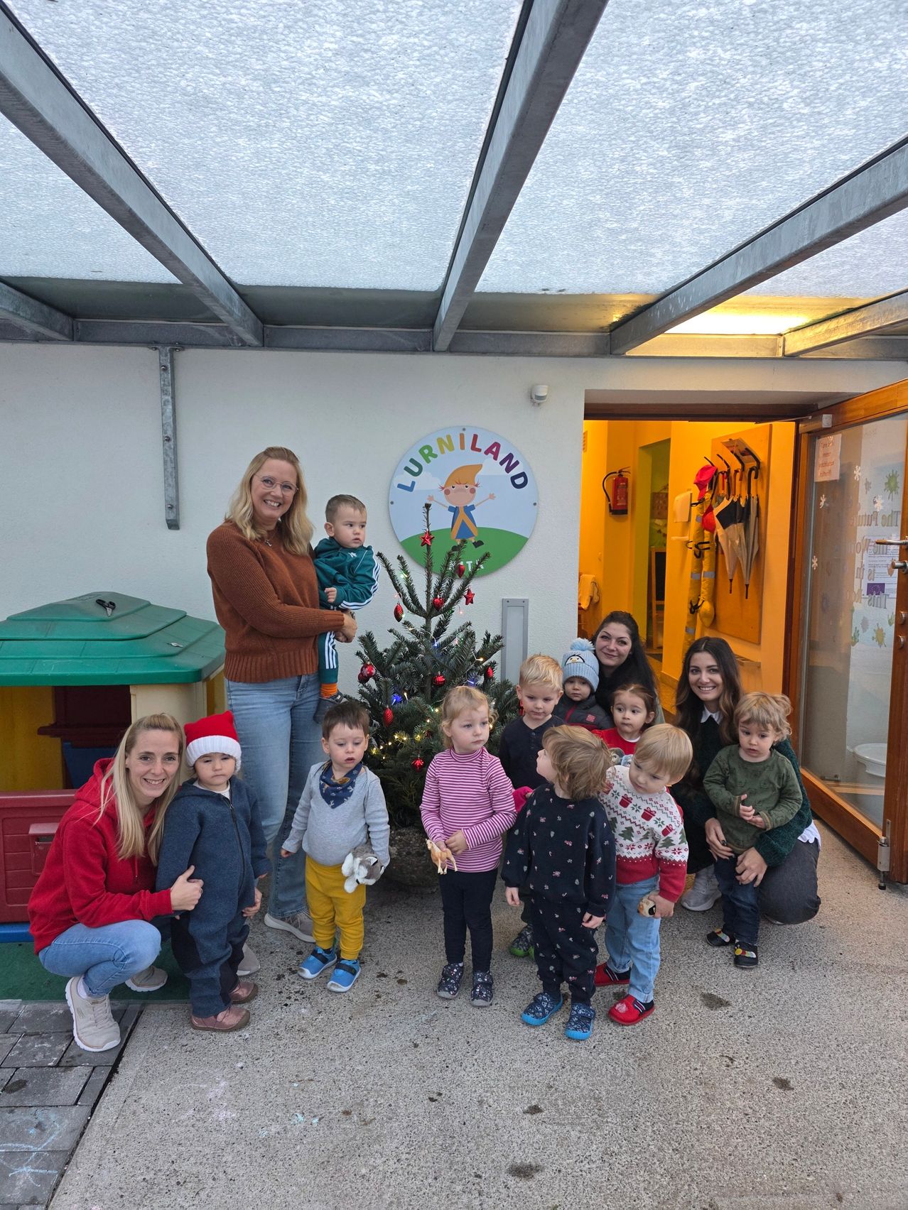Eine Gruppe lächelnder Erwachsener und Kinder posiert vor einem Gebäude mit einem Weihnachtsbaum, einem Schild mit der Aufschrift 'Lurnland' und einem an der Wand montierten Feuerlöscher.