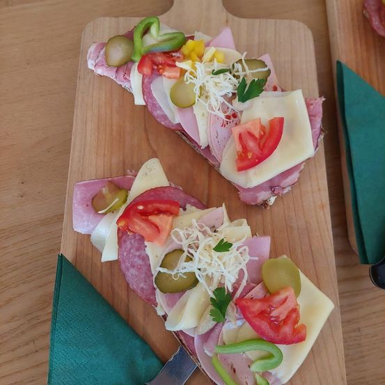 Zwei Scheiben Fleisch- und Käsesandwichs mit verschiedenen Belägen auf einem Holzschneidebrett. Grüne Servietten und ein Messer befinden sich auf der linken Seite.
