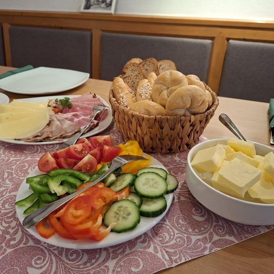 Auf einem Tisch befinden sich verschiedene Speisen: zwei Teller mit Gemüse, Schinken, Käse und Brot in einem Korb sowie eine Schüssel Butter. Stühle umgeben den Tisch.