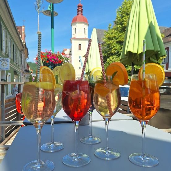 Fünf Weinglas mit farbenfrohen Getränken und Zitronenscheiben auf einem Tisch. Dahinter ein grüner Schirm und ein Turm.