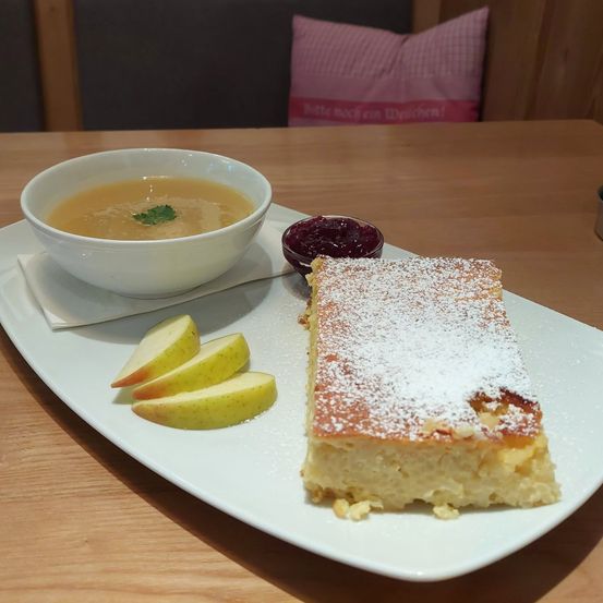 Ein weißer Teller mit Suppe, Apfelscheiben und einem Dessert. Das Dessert hat Puderzucker obenauf.