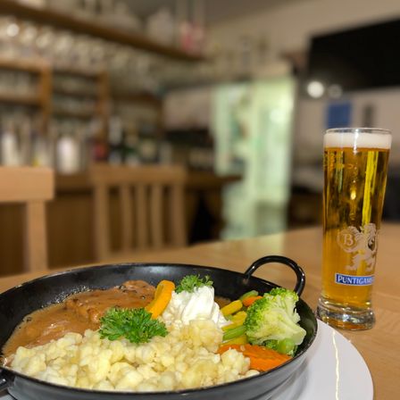 Eine Schüssel mit traditionellem deutschem Essen mit Fleisch, Kartoffeln und Gemüse steht auf einem Tisch mit einem Glas Bier. Im Hintergrund befindet sich eine verschwommene Bar mit Regalen und einem Fernseher.