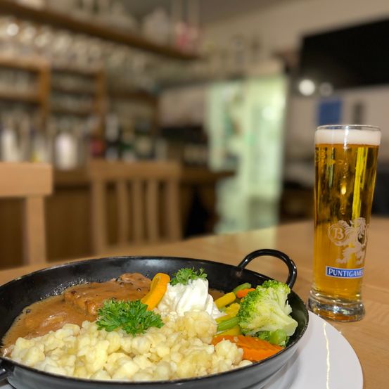 Eine Schüssel mit traditionellem deutschem Essen mit Fleisch, Kartoffeln und Gemüse steht auf einem Tisch mit einem Glas Bier. Im Hintergrund befindet sich eine verschwommene Bar mit Regalen und einem Fernseher.
