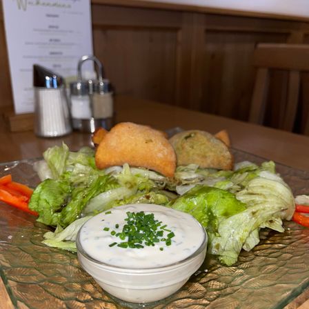 Ein Teller mit Salat, bestehend aus Salatblättern, Karotten und zwei Brotstücken, begleitet von einer Schüssel mit cremiger Kräutersauce. Dahinter befinden sich ein Menü und zwei Salz- und Pfefferstreuer.