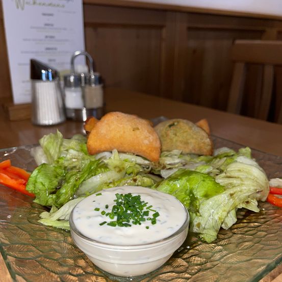 Ein Teller mit Salat, bestehend aus Salatblättern, Karotten und zwei Brotstücken, begleitet von einer Schüssel mit cremiger Kräutersauce. Dahinter befinden sich ein Menü und zwei Salz- und Pfefferstreuer.