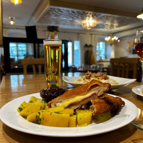 Ein Restauranttisch mit einem Teller gebratenem Schinken-Käse-Sandwich, Kartoffelpüree, Cranberry-Sauce und einem Glas Bier und Wein. Der Hintergrund ist verschwommen mit Stühlen, Tischen und Lampen.