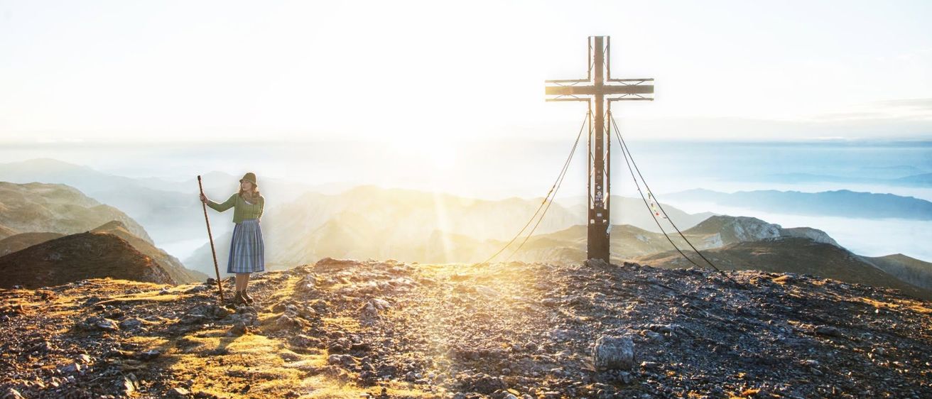 Eine Frau steht auf einem Berggipfel bei Sonnenuntergang, hält einen Wanderstock und hat ein großes Kreuz im Vordergrund.