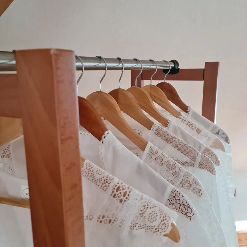 Eine Reihe weißer Blusen mit Spitzendetails ist ordentlich an Holzbügeln aufgehängt und wird an einer Metallstange vor einer schlichten weißen Wand präsentiert.