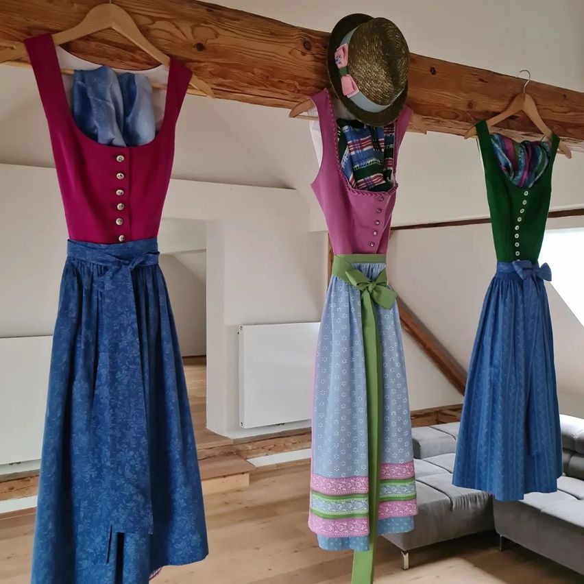 Drei traditionelle Kleider hängen an einem Holzbalken in einem Raum. Jedes Kleid ist in verschiedenen Farben und Stilen, eines hat einen Hut darüber. Der Raum hat Holzböden und weiße Wände.