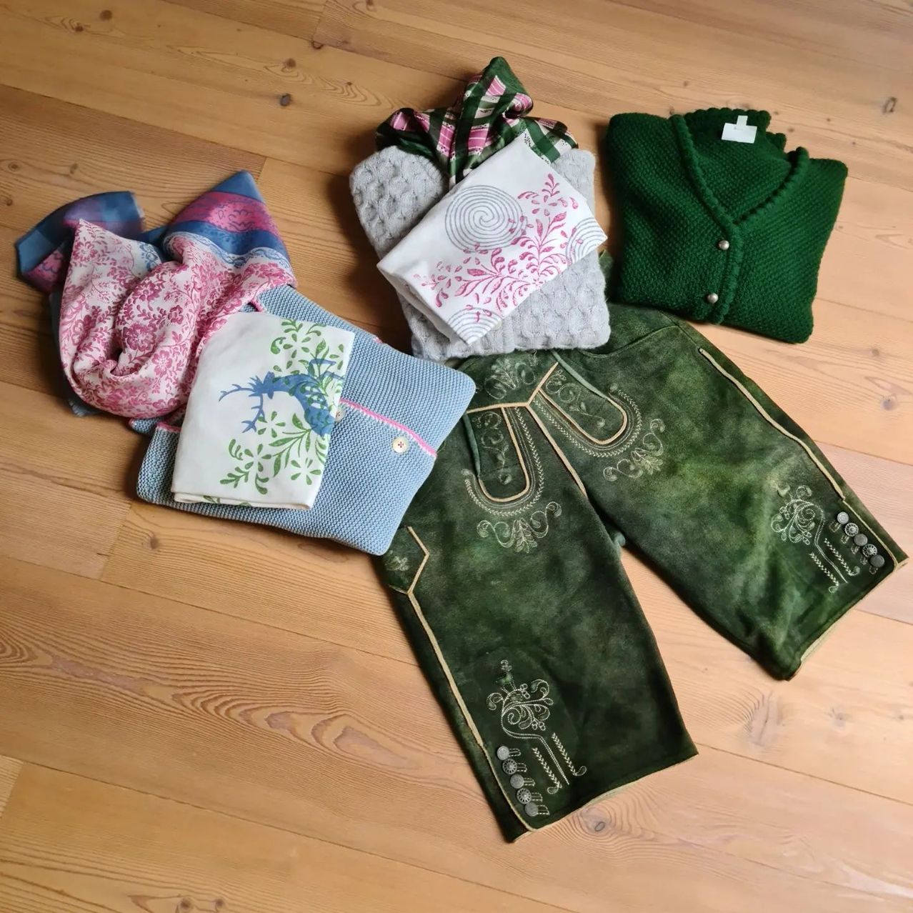 Eine Sammlung von Kleidungsstücken, darunter grüne bestickte Hosen, ein grüner Strickcardigan, ein grauer Schal und ein weißes Taschentuch mit Blumendesigns, sind auf einem Holzboden ausgelegt.