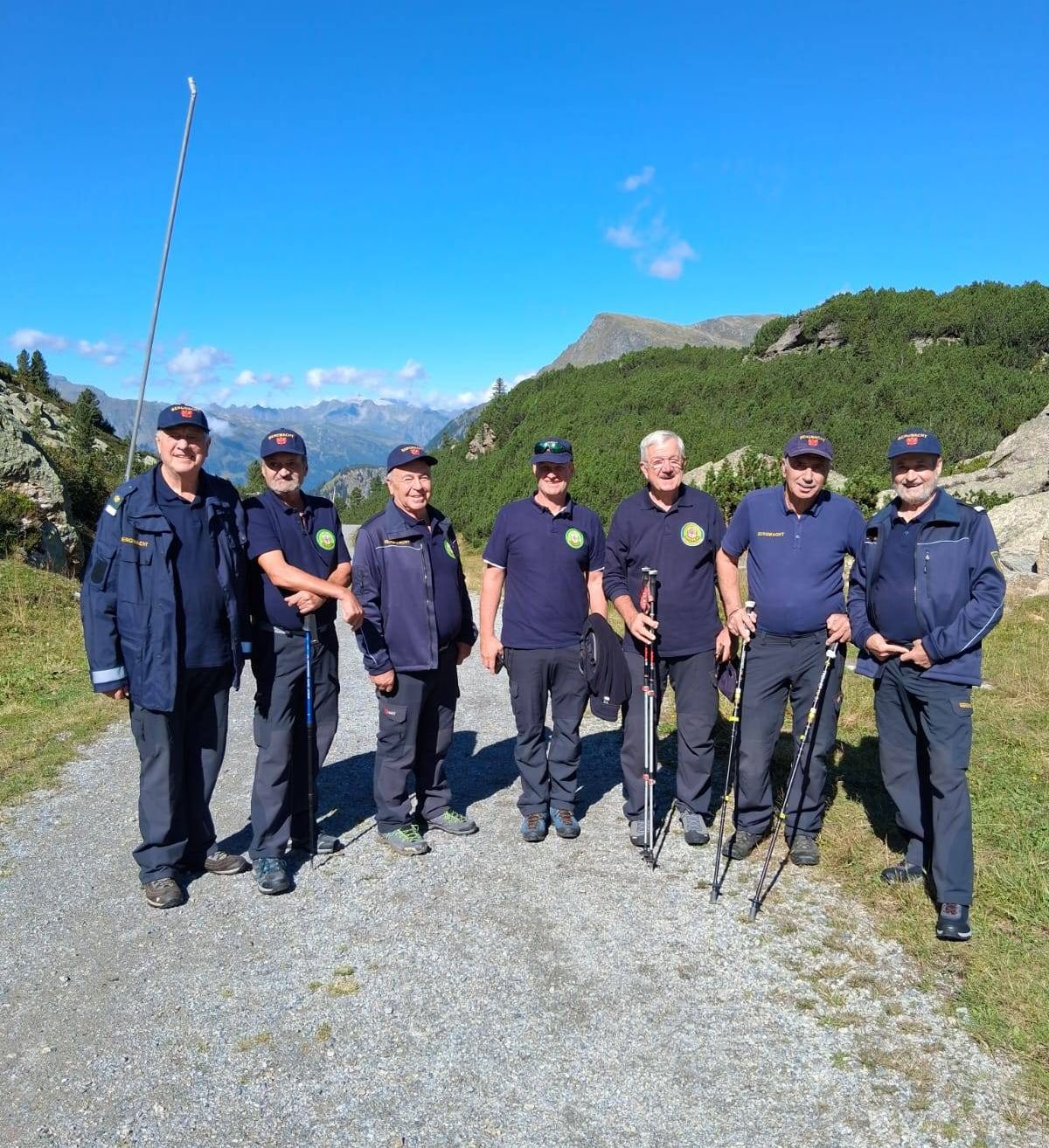 Sieben Männer in blauen Uniformen stehen auf einem Kiesweg und lächeln für ein Foto. Sie halten Trekkingstöcke und sind von einer Berglandschaft umgeben.