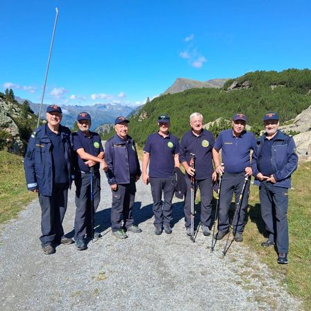 Sieben Männer in blauen Uniformen stehen auf einem Kiesweg und lächeln für ein Foto. Sie halten Trekkingstöcke und sind von einer Berglandschaft umgeben.