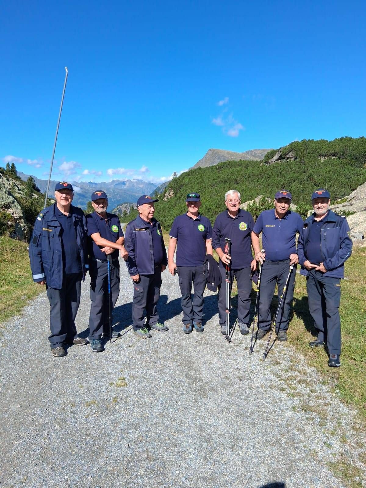 Sechs Männer in identischen Uniformen stehen auf einem Kiesweg, lächeln und halten Trekkingstöcke, mit einem bergigen Hintergrund.