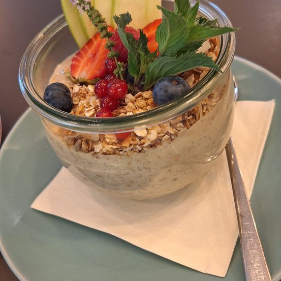 Ein Glas mit Haferbrei mit frischen Beeren, Minze und Granola auf einem grünen Teller mit einem Löffel.
