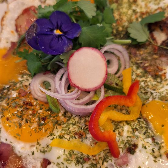 Eine Nahaufnahme eines Gerichts mit gebratenen Eiern, violetten Blumen, geschnittenen Radieschen und lila Zwiebeln. Kräuter und Gewürze sind darüber gestreut.