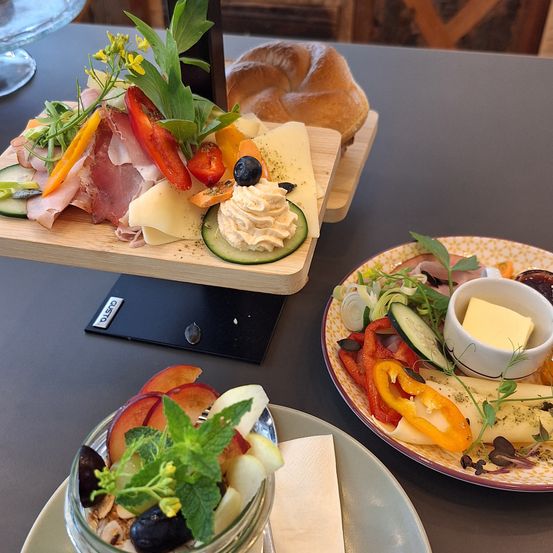 Auf einem Tisch sind verschiedene Speisen zu sehen: eine Käseplatte mit Fleisch und Gemüse, ein Obstsalat und ein Teller mit Käse und roten Paprikaschoten.