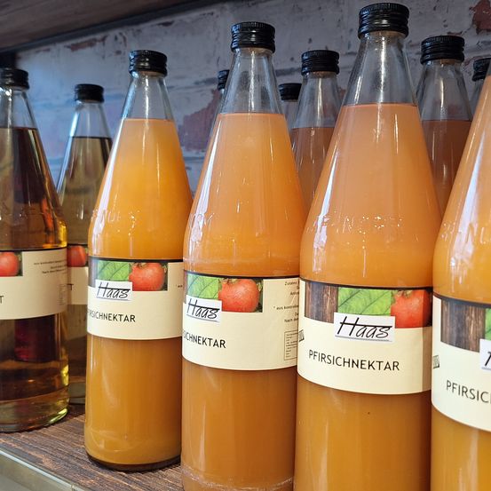 Mehrere Saftflaschen sind auf einem Holzregal aufgereiht. Die Flaschen sind mit orangefarbener Flüssigkeit gefüllt und haben Etiketten mit den Worten 'Haas' und 'Pfirsichnektar' darauf.