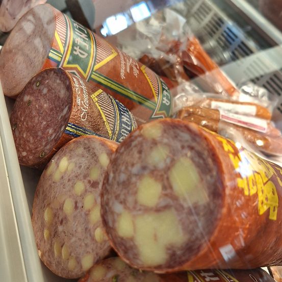 Eine Glasvitrine zeigt verschiedene Würste. Einige sind in Plastik verpackt, andere in ihrer Originalverpackung. Die größte Rolle ist aufgeschnitten und zeigt einen Querschnitt mit sichtbaren gelben Stücken.
