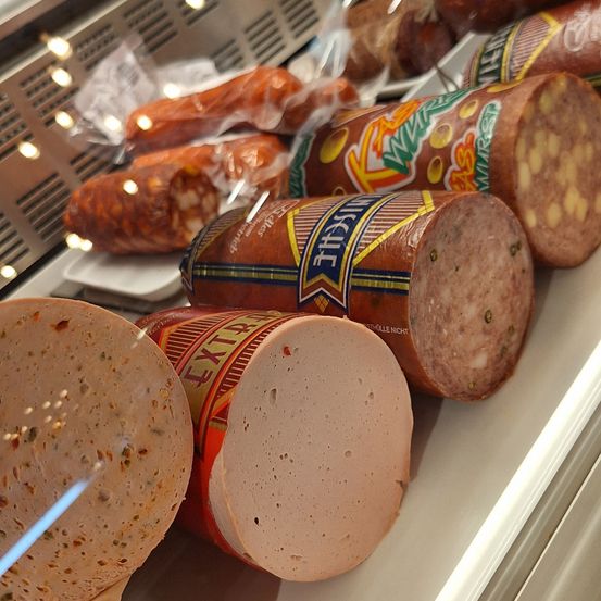 Eine Vitrine zeigt verschiedene Wurstsorten, darunter eingewickelter Salami, eine große Wurst mit der Aufschrift 'Mettwurst' und eine runde Wurst in einer Plastikverpackung mit einem rot-gelben Etikett.