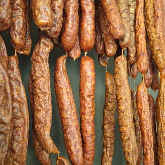 Eine Sammlung von geräucherten Würsten, wahrscheinlich Chorizo, hängt an einer Wand. Die Würste haben eine rotbraune Haut mit einer leicht welligen Textur, die von dünnen Schnüren hängen. Die Würste sind in einer Reihe angeordnet, wobei der oberste Wurst der größte ist.