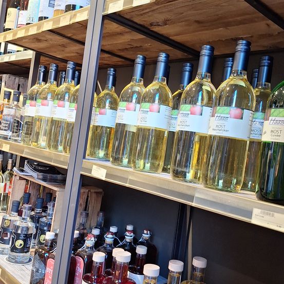 Ein Ladenregal mit einer Reihe von Flaschen mit klarer Flüssigkeit und blauen Verschlüssen, gekennzeichnet als Apfelwein. Darunter ein weiteres Regal mit verschiedenen Spirituosenflaschen, einige mit weißen Verschlüssen.