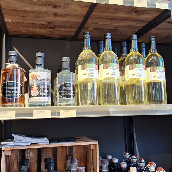 Ein Holzregal zeigt mehrere Flaschen mit weißen Etiketten und Text. Darunter befindet sich ein hölzerner Kasten mit Papieren.