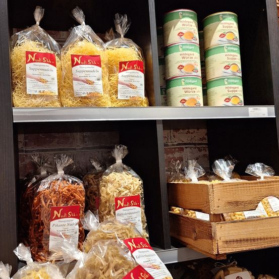 Ein Regal mit verschiedenen Nudel- und Suppenprodukten. Verschiedene Nudelarten sind in Plastiktüten verpackt, und Dosensuppe ist auf dem oberen Regal gestapelt. Holzkisten mit Nudeln stehen auf den unteren Regalen.