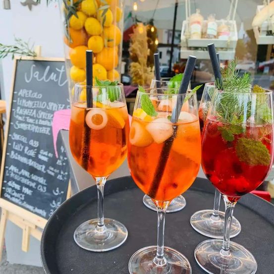 Drei hohe Gläser mit verschiedenen Cocktails, Strohhalmen und Garnierungen stehen auf einem Tablett vor einer Kreidetafel-Menü.