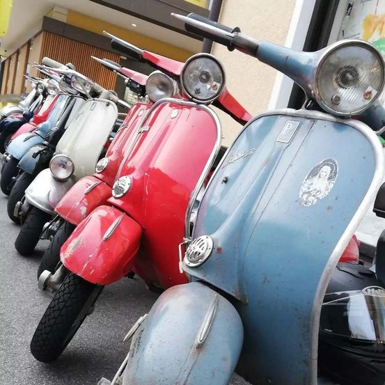 Eine Reihe von Oldtimer-Rollern ist vor einem Gebäude geparkt. Sie sind in verschiedenen Farben, darunter rot, weiß und blau. Die Roller haben Aufkleber und einige haben einen Helm auf dem Sitz.