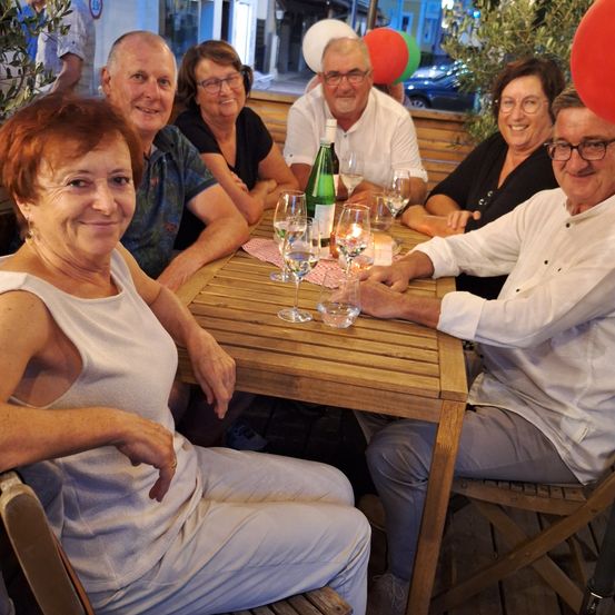 Eine Gruppe von Menschen sitzt draußen an einem Tisch und lächelt für ein Foto. Sie haben Weingläser und eine Flasche Wein auf dem Tisch. Hinter ihnen befinden sich Luftballons und ein Auto.