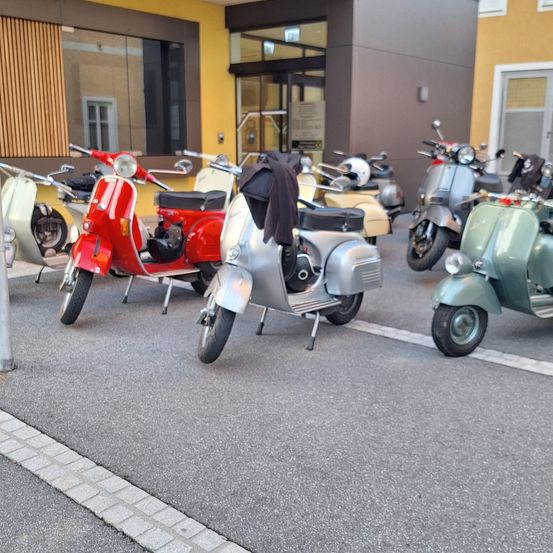Mehrere Vintage-Roller sind vor einem Gebäude geparkt, einige mit Jacken auf den Sitzen.