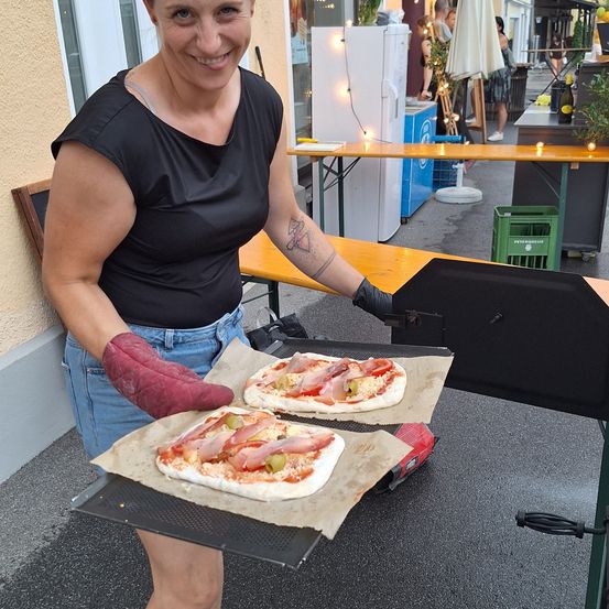 Eine Frau in einem schwarzen Shirt und roten Ofenhandschuhen hält zwei Pizzen auf einem Tablett draußen. Hinter ihr sind ein Kühlschrank und ein Tisch mit einer Topfpflanze zu sehen.