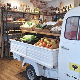 Ein weißer Lebensmittellastwagen mit einer Auswahl an frischem Gemüse und Früchten in Körben. Hinter dem Lastwagen befinden sich Regale mit Flaschen Wein und anderen Getränken.