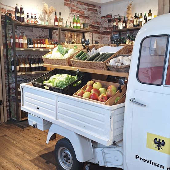 Ein weißer Lebensmittellastwagen mit einer Auswahl an frischem Gemüse und Früchten in Körben. Hinter dem Lastwagen befinden sich Regale mit Flaschen Wein und anderen Getränken.