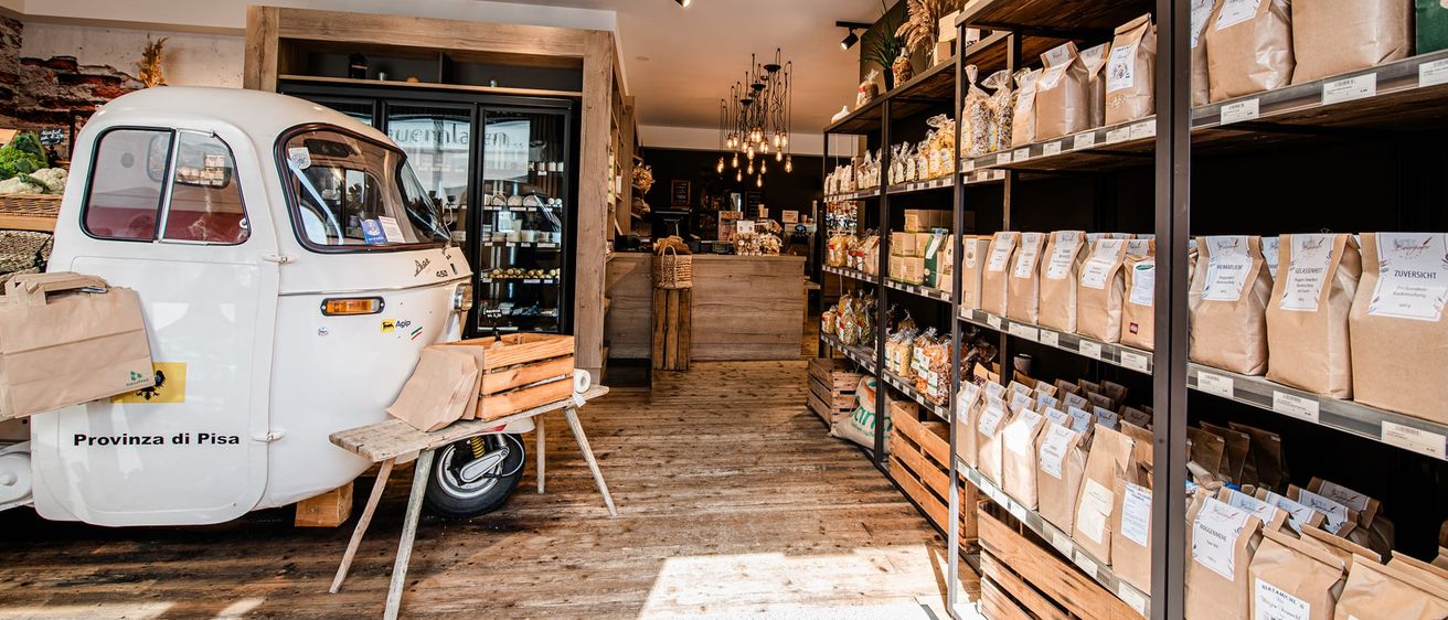 Innenansicht eines Geschäfts mit Holzböden und Regalen. Säcke mit verschiedenen Artikeln sind auf den Regalen ausgestellt. Ein weißes Fahrzeug ist in der Nähe einer Holzbank geparkt.