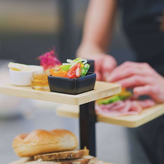 Ein Nahaufnahme eines hölzernen Etagentabletts mit verschiedenen Lebensmitteln, einschließlich einer Schüssel mit Gemüse, einem kleinen Glas mit Senf und Fleischscheiben, mit den Händen einer Person im Hintergrund.