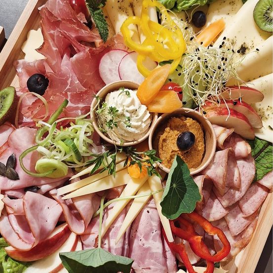 Ein Holztablett zeigt eine Auswahl an Fleisch, Käse, Gemüse und Obst. Geschnittenes Fleisch ist neben Käsestücken und verschiedenen Gemüsesorten angeordnet. Schüsseln mit Aufstrichen und Früchten sind enthalten.
